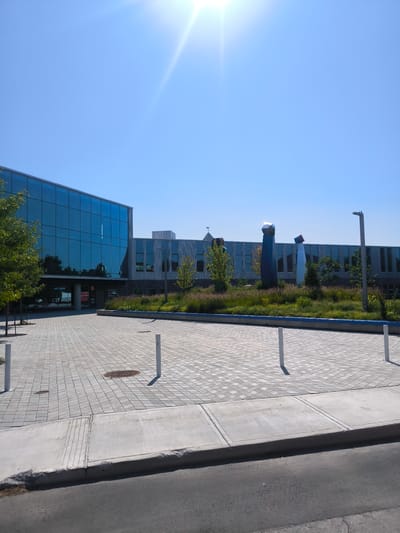 This is a horizontal, eye-level shot of a modern building with a glass facade and a landscaped area in front. The sun is shining brightly in a clear blue sky, indicating it is a sunny day, likely mid-morning to early afternoon.
The building has a distinctive design, with a large section featuring a wall of blue-tinted glass panels, reflecting the sky and surrounding trees. Adjacent to this is a section with a corrugated metal facade and rectangular windows. In front of the building, there is a paved plaza with a brick-like pattern, bordered by a curb and a road. White bollards are strategically placed throughout the plaza.
The landscaped area features a variety of grasses and greenery, with a vibrant blue border along its edge. Three tall, abstract sculptures are prominently displayed in the landscaped section. These sculptures appear to be made of reflective material, with geometric shapes at their peaks that catch the sunlight. One sculpture is dark blue, another is a lighter blue or silver, and the third is a reddish-brown. A weather vane is visible atop the building in the distance.
There are no people visible in the image. The scene appears to be a public space or an entrance to a commercial or institutional building, possibly in Chambly, Canada, given the location context. The overall impression is one of a modern, well-maintained, and aesthetically pleasing environment.