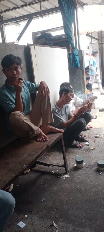 The image depicts a semi-enclosed, sheltered area in Cilampeni, Indonesia, occupied by a group of men. In the foreground-left, a male subject in a light blue polo shirt and khaki pants sits barefoot on a worn wooden bench, smoking a cigarette with visible ash. He rests his right arm on his raised right knee, looking towards the right side of the frame. Adjacent to him, towards the mid-ground right, another male subject wearing a grey t-shirt and dark pants with black sandals squats on the concrete ground, attentively viewing a mobile phone held in both hands. Behind this individual, a third male in a white t-shirt is partially visible, also squatting or sitting, with a plume of smoke indicative of smoking.

The ground surface is dark, likely concrete or packed dirt, and appears unkempt with scattered litter including cigarette butts, paper, and small plastic debris. A clear plastic bottle with a red cap and two small cylindrical containers with dark green lids are present on the ground. The background features a light-colored wall or partition, and beyond that, an open area where a fourth male subject in a light blue shirt and white pants is seen walking away from the camera. Overhead, a corrugated metal roof structure with visible beams provides shelter. A length of blue fabric is suspended from an overhead beam. Natural light illuminates the scene, originating primarily from the upper right and background.