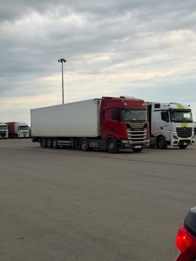The image shows several parked semi-trailer trucks on a paved surface, possibly a parking lot or a loading area. The trucks are lined up side-by-side. The sky is cloudy, suggesting overcast weather, and the lighting indicates it is daytime, possibly late afternoon or early evening given the softness of the light.

The most prominent truck is a red Scania with a white trailer. Another truck visible is a white Mercedes-Benz with yellow and green accents on its cab, also pulling a white trailer. Several other trucks are partially visible behind these. The trucks appear to be stationary, suggesting they are parked, possibly waiting for loading or unloading.

In the background, a tall light pole with multiple lights is visible against the cloudy sky. The overall scene is utilitarian, characteristic of a logistics hub or industrial area. There is no visible indication of people in the image.

Visible text includes "SCANIA SUPER" on the grille of the red truck. The license plate on the red truck appears to be "C438MP 152". The white Mercedes-Benz has text on its side, with "ТЕХНОЛОГИЯ" and "ПРОДВИЖЕНИЯ" being partially legible.

The location is identified as Усть-Сыны, Russia.