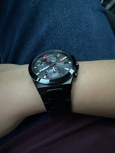 A close-up shot features a black Casio Edifice solar-powered watch on a person's wrist. The watch has a dark grey face with silver hands and markers, and three smaller subdials. The band is also black and appears to be made of metal. The wrist is positioned against a background of dark blue denim fabric, likely from jeans, and a darker, possibly maroon, textured fabric, likely from a shirt or pants. The lighting suggests an indoor environment, possibly with ambient light. The image does not contain any identifiable landmarks or features that would indicate its location within the city of Saedinenie, Bulgaria.