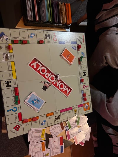An overhead view captures a Greek Monopoly board game in progress on a light brown tabletop in Gerakas Municipal Unit, Greece. The board prominently features the red "MONOPOLY" logo diagonally across its center. Two white dice, showing faces of four and three dots, rest near a black top hat game token. A silver car token and a silver thimble token occupy property spaces on the left side of the board. Red houses and green houses are distributed across various property spaces: three red houses on an orange set, two green houses on a light blue space, four red houses on a top-side light blue set, and three red houses with two green houses on a dark pink set. A stack of orange Chance cards with a question mark is face-down on the top-right quadrant of the board, while a stack of light blue Community Chest cards with a chest icon is on the bottom-left quadrant. At the top of the image, a black plastic money tray contains sorted Monopoly money in denominations of 1, 10, 20, 50, 100, and 500. Below the board, numerous property deed cards of various colors are fanned out and scattered across the table. The lower half of an individual's legs, clad in light grey sweatpants, is visible on the right, suggesting a player seated at the table. A dark clothing item is partially visible on the far left, indicating at least one other participant. The table rests on a darker, possibly tiled floor.