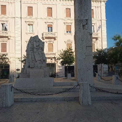 The image captures an urban scene in Piazza Garibaldi, Pescara, Italy, under a clear blue sky. In the mid-ground, two distinct stone monuments are visible. To the left, the Monumento ai Caduti (War Memorial) is a prominent, large, rough-hewn stone structure with a jagged, naturalistic form, set upon a multi-tiered plinth of smoother, lighter-colored stone steps. To the right, partially cropped, stands a tall, rectangular column, identified as a component of La Nave di Cascella monument. This column is constructed from large, interlocking light-colored stone blocks, some featuring puzzle-piece-like shapes.

The foreground consists of a light-colored, textured paved ground. Several hexagonal stone bollards are positioned around the base of the monuments, connected by dark metal chains, forming a perimeter barrier. No individuals are present within the frame.

In the background, a multi-story building with a light pink or salmon-colored facade dominates. This building features numerous windows, many fitted with closed light brown external shutters. White architectural elements, including window frames, cornices, and decorative balcony balustrades, adorn the facade. Laundry, specifically a white garment, is visibly hanging from a balcony on the far left of the building. A white banner displaying a blue logo is affixed to the upper right section of the building's facade.

Several small to medium-sized green trees are interspersed between the monuments and the background building. A black bicycle is parked near a lamppost to the left of the Monumento ai Caduti.