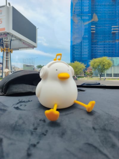 The image depicts a dashboard view from within a vehicle. In the foreground, a white duck figurine with yellow feet and a yellow musical note on its head is positioned centrally. To the left of the figurine is a black baseball cap. The background reveals a street scene with trees, a billboard, and a tall building under construction covered in blue netting. The setting is likely An Nhơn, Vietnam, based on the architectural style and visible signage. The image appears to be taken during daylight hours.