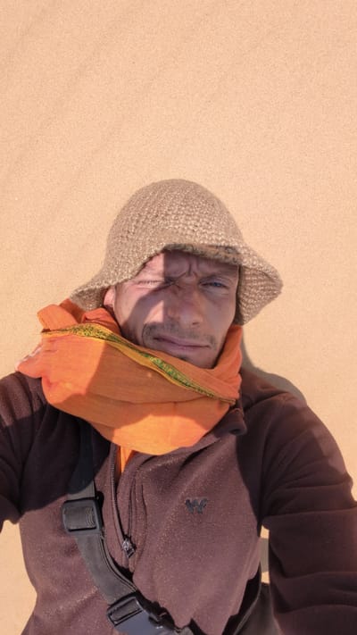 A man lies on a sandy surface, likely a beach, in Phuoc Dinh, Vietnam. He is wearing a brown fleece jacket with a black strap across his chest, an orange scarf around his head, and a beige knit cap. His face is partially in shadow. The sand is a uniform tan color.