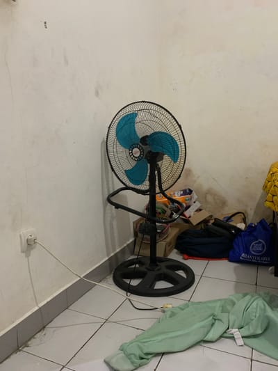 The image captures an indoor scene in Cibinong, Indonesia, depicting a cluttered corner of a room. The primary subject is a standing floor fan with a black metal cage and bright blue blades, positioned prominently on the left side of the frame. The fan's central hub bears partially visible text, possibly "OKAY".
The room's walls are a light, possibly off-white, color and display significant signs of wear and tear, including numerous stains, dirt marks, and visible cracks, particularly near the base and extending across the surfaces. The floor is covered with large, light-colored square tiles that also show grime and some cracks, especially along the edges and grout lines.
In the lower right foreground, a white power outlet is installed on the wall, with a white power cord plugged into it. This cord extends towards the right, disappearing from view, suggesting it powers the fan or another nearby appliance.
To the right of the fan, the corner of the room is filled with various stored items. These include several cardboard boxes, some stacked and appearing either empty or containing miscellaneous goods. Piles of fabric are also visible, most notably a light green sheet or piece of clothing draped on the right, and a patterned yellow fabric near the top edge. A blue bag, possibly a reusable shopping bag or a uniform, is perched atop other items, displaying the white text "BHAKTI KARYA" and "BERSAMA KITA BERKARYA". A dark, circular object, resembling a weight plate, is also discernible amongst the clutter.
No people are present in the image, and no specific activity is taking place. The lighting suggests an interior setting, likely illuminated by ambient daylight or artificial light sources. The overall impression is that of a utility space, storage area, or a general room that is not meticulously maintained.