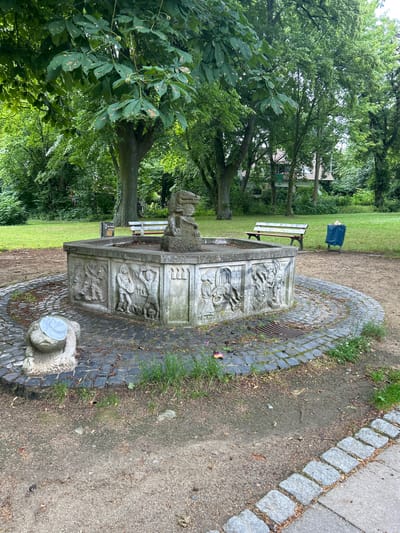 This media file captures a quaint park scene in Königslutter am Elm, Germany. The primary subject is an ornate, old stone fountain with intricate carvings depicting what appear to be figures and mythical creatures. The fountain is situated on a circular cobblestone base surrounded by a gravel path and lush green grass.

In the background, a white park bench is visible, with a dark blue trash bag resting on it, suggesting the area is maintained for public use. Several large trees with dense green foliage frame the scene, creating a shaded and natural environment. A building with a tiled roof is partially visible through the trees in the distance.

The image appears to be taken on a bright, possibly overcast day, given the even lighting and the absence of strong shadows. The overall mood is peaceful and serene, characteristic of a quiet park setting. There is no visible text in the image. The time of day is likely daytime, given the natural light. The presence of well-maintained greenery and public amenities like the bench and fountain points to a developed park area. The carvings on the fountain suggest historical or artistic significance to the location.