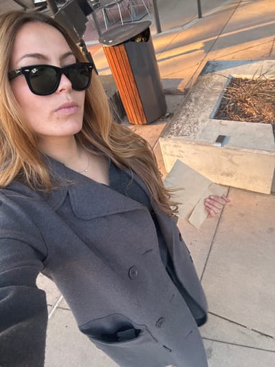 A woman takes a selfie outdoors on a sidewalk. She wears sunglasses and a gray overcoat. Her hair is long and blonde. She holds a brown paper bag in one hand. A planter box and a trash receptacle are visible in the background, along with pavement and building shadows. The photo has a slightly tilted perspective.