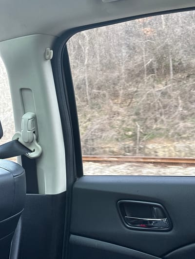 The image depicts a view from the interior of a vehicle, looking out a passenger-side window in Leshnitsa, Bulgaria. The foreground on the left features a light-colored B-pillar with an attached seatbelt height adjuster and a black seatbelt strap. A portion of a black vehicle seat is visible in the lower left. The lower right foreground shows a dark, textured door panel with a chrome door handle that includes a small red reflective element.
Through the window, an outdoor environment is visible. Horizontally across the lower half of the frame, a set of railroad tracks with two rusty-brown rails on grey ballast runs parallel to the vehicle's direction of travel. Beyond the tracks, a natural slope rises, covered with numerous bare, leafless deciduous trees and sparse dry vegetation, suggesting a winter or early spring season. The window glass exhibits a speckled appearance, possibly from dirt or moisture. No individuals or specific actions are discernible within the frame.