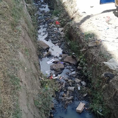 The image displays an open, heavily polluted drainage channel or stream within Ongata Rongai ward, Kenya. The channel, estimated at 2-3 meters wide, features steep earthen banks partially covered by dry, sparse grass and limited green vegetation. The water within the channel is dark and appears stagnant, heavily laden with refuse. Identifiable objects include numerous plastic bottles (clear, brown, green), various plastic bags (red, white, black), paper debris, and fragments of cardboard, including a distinct brown cardboard box floating. Large rocks and dark, unidentifiable debris are interspersed throughout the watercourse. On the right bank, a single discarded sandal is visible. In the upper right quadrant, a partial blue object, consistent with a tarp or vehicle component, is observed. The scene is illuminated by direct daylight, casting shadows on the right bank. No individuals, actions, or interactions are depicted.