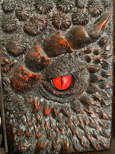 This is a close-up, overhead shot of a carved, textured object, possibly a decorative box or a book cover, with a dragon or monster-like design. The primary subject is the textured surface itself, featuring a prominent, realistic-looking red eye as the focal point. The eye is rendered with a glossy finish, reflecting light and giving it a lifelike appearance.

The surrounding texture is complex, with various raised and indented areas that suggest scales, spikes, or rough skin. The color palette is dominated by dark, metallic grays and blacks, heavily accented with vibrant streaks of fiery orange and red, particularly around the eye and along the edges of the raised textures. This coloration gives the impression of heat, danger, or embers.

The texture is highly detailed, with fine lines and rough surfaces contributing to a tactile, almost organic feel. Some areas exhibit starburst-like patterns, perhaps representing smaller scales or natural formations. The lighting is harsh, creating strong shadows that emphasize the three-dimensionality of the carving.

There is no discernible human presence, activity, or event taking place, as the image focuses solely on the static, decorative object. The time of day, weather, and location cues are not apparent from the image itself, other than the provided context of "Alamüdün, Kyrgyzstan," which refers to the origin of the image file, not a visible element within the image. There is no visible text in the image. The overall impression conveyed is one of fierce, mythical power and intricate craftsmanship.