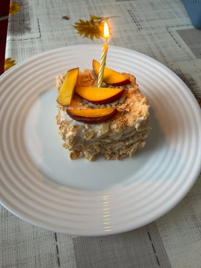 A slice of layered cake, likely Napoleon or Mille-feuille, sits on a white, ridged plate. The cake is topped with several slices of fresh peach and one lit birthday candle with a yellow and white striped pattern. The plate is placed on a tablecloth with a subtle checkered pattern and floral accents. The scene is indoors, likely on a table, with the lighting suggesting it could be daytime. No specific location in Holíč, Slovakia is discernible from the image.