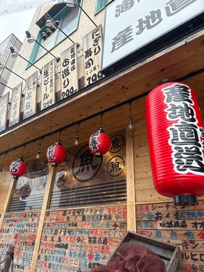The image depicts a building in Takarazuka, Japan, with coordinates 34.81, 135.345. The building is constructed of wood and has a sign that reads "Takarazuka" in Japanese. There are several red paper lanterns hanging in front of the building, which are lit. The building has a window with a display of food and drink inside. The building also has several signs in Japanese that are advertising various products. The signs are written in black paint. The building is located on a street, and the street is lined with other buildings and businesses. There are no people visible. The weather appears to be overcast with light rain.