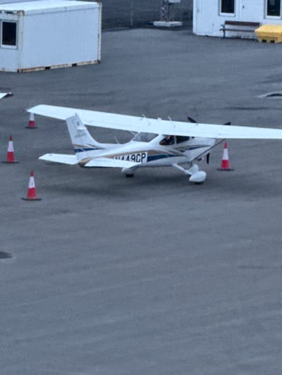 The image shows a white and blue single-engine airplane parked on an asphalt surface, likely an airport tarmac. The aircraft has the registration number "N449CP" visible on its tail. Three orange and white traffic cones are positioned around the plane, suggesting it is in a designated parking or movement area. In the background, there is a white modular building, resembling a shipping container, with a dark window. Further in the background, a white building with multiple windows and a small yellow object are visible, possibly part of airport infrastructure. The overall scene is overcast, with a muted color palette typical of a cloudy day. This scene is consistent with the area around Reykjavik City Airport (RKV) in Reykjavik, Iceland, known for its general aviation activities.