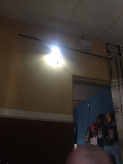 The image displays an interior room located in Oba-Ile, Nigeria. The visible wall is painted in two horizontal sections: an upper pale yellow and a lower dark brown, separated by a thin white stripe. A single, intensely bright, unshielded light source is affixed to the upper left portion of the pale yellow wall, generating significant glare. A dark, thin horizontal bar is mounted across the upper wall section. Near the ceiling on the right, a circular, flush-mounted light fixture is present. An open doorway to the right leads into an adjacent room, revealing a distinct blue wall adorned with multiple hanging objects, including hats, bags, and framed items. A patterned curtain with a dark border partially obscures the view into this adjacent room. The ceiling is light-colored with visible structural beams. The immediate foreground is blurred and dark, without discernible features. No individuals or specific actions are identifiable within the scene.