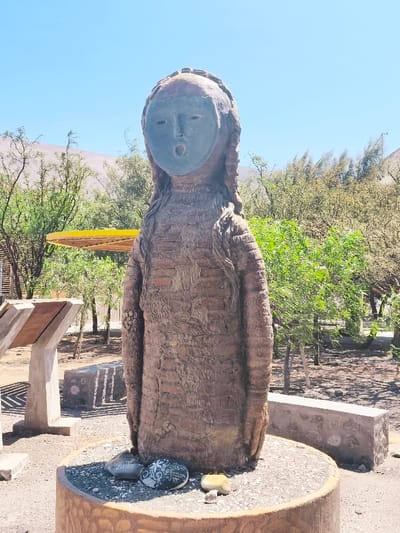 The image displays a weathered anthropomorphic statue centrally positioned in an outdoor setting in Camarones, Chile. The statue, standing erect, is crafted from a reddish-brown material, presenting a textured surface akin to rough fabric or bark, with horizontal striations visible across its body and braided hair details. A smooth, dark grey-blue mask covers the face, featuring empty eye sockets and a distinct circular opening for the mouth. The statue is anchored to a cylindrical, light brown concrete base topped with small gravel, upon which several smooth, small stones, some bearing inscriptions or patterns, are meticulously arranged.

In the midground behind the statue, sparse yet vibrant green and lighter brown arid vegetation, including trees and shrubs, are present. To the left, a concrete informational display stand with a tilted brown panel is visible, along with a partial view of a yellow, circular or dish-shaped structure mounted on a pole, partially obscured by foliage. Scattered concrete benches or low walls with stone-textured finishes are also discernible. The immediate ground surface consists of light brown, dry earth or gravel. The background reveals light brown, arid mountains or hills under a clear, bright blue sky, indicating strong daylight.