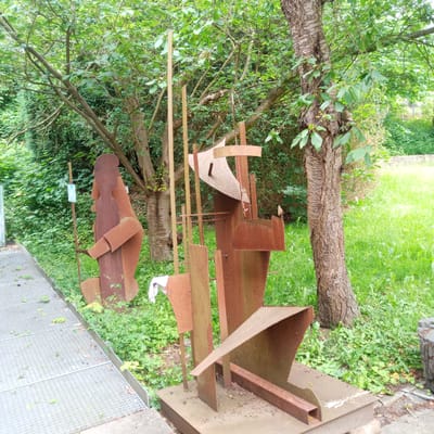 The image shows two abstract metal sculptures in an outdoor setting, with lush green trees and grass. To the left, a dark brown, roughly humanoid figure is positioned behind a metal pole with a blue sign. To the right, a more complex, rusty metal sculpture with angular shapes and curved elements is situated. A large tree trunk is visible behind the sculptures, and a pathway made of metal grating runs along the left side of the scene. The ground is covered in green vegetation and some fallen leaves. The scene appears to be a public space, possibly a park or sculpture garden, in the city of Eisenach, Germany.