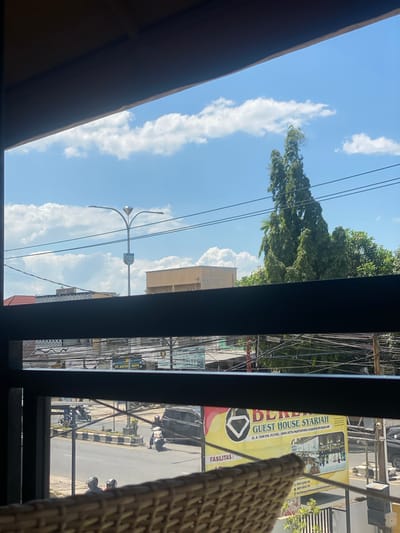 The image depicts a street scene viewed through a window, with a dark, horizontal bar in the foreground. The sky is blue with scattered white clouds. In the middle ground, a tall, slender evergreen tree stands prominently. Below it, a street is visible with two motorcycles and a car. A yellow billboard for "BERLIAN GUEST HOUSE SYARIAH" with an address in Martapura, Banjar Regency, Indonesia, is also present. Utility poles and electrical wires are strung across the scene, suggesting an urban environment. The overall impression is that of a sunny day in a populated area.