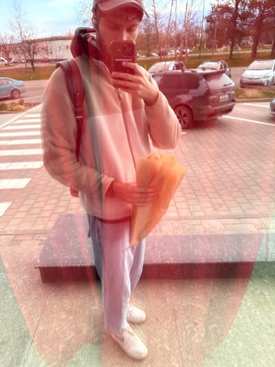 A person is taking a selfie in front of a building. The building is likely located in Ciorescu, Moldova. The person is wearing a beige jacket, blue jeans, and white sneakers. They are holding a brown paper bag. Cars and trees are visible in the background, reflected in the glass that the person is standing next to. There is a crosswalk near the person.