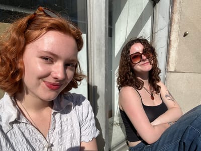 The image features two young women lying side-by-side on a light-colored outdoor surface, likely concrete or pavement, under bright sunshine. They appear to be looking towards the camera, possibly posing for a casual photograph.
The woman on the left has dark, curly hair and is wearing dark sunglasses. She is dressed in a black spaghetti strap top and blue denim fabric, possibly trousers or a skirt, with her arms crossed over her chest. A tattoo of a delicate branch or floral design is visible on her left forearm. Around her neck, she wears a pendant necklace with a reddish-brown stone. Her expression is relaxed.
The woman on the right has reddish-brown hair. She wears a light-colored, possibly white or light grey, textured top with a shirred or smocked design and a collared neckline. A silver chain necklace is visible, and sunglasses with tortoiseshell frames rest on her head. Her lips are slightly parted, revealing a reddish tint, and she also displays a relaxed expression.
Strong sunlight casts prominent shadows, indicating a clear, sunny daytime. A vertical light-colored structure, possibly a window frame or building facade, runs between the two individuals. A small, metallic, bullet-shaped object is visible on the concrete surface near the left woman's head. The scene conveys a relaxed outdoor moment in Dundee, United Kingdom. No text is visible in the image.