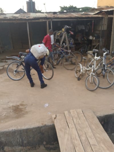 The image captures an outdoor scene in what appears to be a bicycle repair shop or market stall in Ijaiye, Nigeria. The setting is rustic, with a structure made of corrugated metal sheets serving as the shop, visible in the background.
In the foreground, a person wearing blue overalls or pants and a white backpack is bent over a bicycle, seemingly working on it or inspecting it. To their right, another man in a red and white shirt stands near the entrance of the shop, with several bicycles stacked or leaning behind him.
Numerous bicycles are scattered across the dirt ground and within the shop area. Some are parked upright, others are leaning against each other, and several appear to be in various states of repair or display. A small, white children's bicycle is also visible among the adult bikes on the right side of the frame.
The ground is unpaved dirt, and a small wooden plank bridge or ramp extends from the lower right corner, crossing what might be a shallow ditch or drain. In the distance, above the shop's roof, a dark water tank on a stand is visible, along with some sparse vegetation. The lighting suggests it is daytime, possibly overcast or late afternoon, with no harsh shadows. No discernible text is visible in the image.