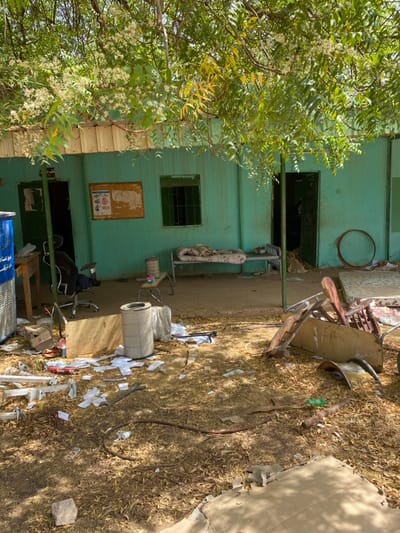 The scene is an exterior view of a building located at the coordinates 15.552 latitude and 32.57 longitude in Khartoum, Sudan. The building is a single-story structure with a green exterior and a roof that is partially obscured by a leafy tree. The building has multiple doors and windows, including one with a barred window and a metal door. The building appears to be abandoned, with debris scattered around the yard. There are numerous pieces of broken furniture and other objects. There are no visible people in the scene. The environment is dry and dusty with scattered leaves on the ground. The image was taken on a bright, sunny day. The time of day cannot be determined from the image.