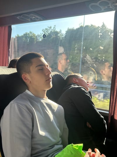 Two young men are sleeping on a bus. The man in the foreground is wearing a light gray sweater and has short, dark hair. He is leaning against the seat. Behind him, a man in a dark hoodie is also sleeping with his arms crossed. The reflection of the men in the bus window shows the trees outside. The bus window has a sticker that shows the handicapped seat symbol. The picture was taken during the daytime with a lot of sunlight coming through the window. The location is likely the Municipal Unit of Kato Olympos, Greece.