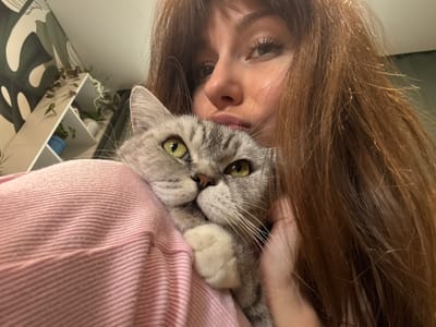 A medium shot captures a young woman with long, auburn hair embracing a gray tabby cat. The woman, facing the camera with a slight upward tilt of her head, has her left eye partially obscured by her hair. Her lips are slightly parted. The cat, held close to her chest and shoulder, looks directly at the camera with bright green eyes. Its front paws are resting on the woman's shoulder, and its tail is visible behind its back. The woman is wearing a pink, ribbed garment, possibly a sweater or shirt. In the background, a white shelf unit is partially visible, holding several small potted plants and adorned with a large, abstract-patterned wallpaper featuring dark green and white shapes. The scene appears to be indoors, in a domestic setting. The photograph was taken in Chaikovsky, Russia.