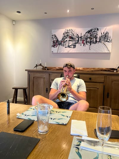 A single adult male is seated indoors, positioned at a light brown wooden dining table. He wears a grey baseball cap, a light-colored t-shirt, and dark shorts, and is actively playing or posing with a golden-colored trumpet held to his lips. On the table in the foreground are a black vape pen, a black smartphone, a clear glass tumbler with white residue, a small white bowl, a folded white napkin, and a set of silverware with a champagne flute, all resting on patterned placemats. In the background, a dark wooden sideboard is visible, topped with a gnarled piece of wood holding three white candles. Above the sideboard hangs a large rectangular artwork depicting a monochrome urban street scene with red accents. A small dark wooden stool is to the left of the man, and a white security sensor is mounted on the far wall. The room is well-lit, featuring light-colored walls and a white ceiling with recessed lighting. The scene is located within the city of Hornsea, United Kingdom.