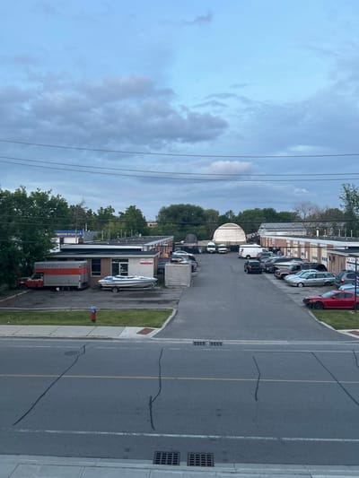 The image depicts a wide view of an industrial or commercial area, likely a business park, under a cloudy sky. In the foreground, a paved road with yellow lane markings and black sealant repairs is visible. A sidewalk and a grassy median with a red fire hydrant separate the road from a parking lot.

The parking lot is lined with several low-rise commercial buildings. To the left, a U-Haul truck is parked in front of a building with a sign that reads "AUTOMOTIVE STATION". Next to the U-Haul truck, a white boat on a trailer is parked.

Further into the lot, a variety of vehicles are parked, including cars, vans, and SUVs. One building appears to be a series of garage bays. In the background, a large, white, dome-shaped structure is prominent, possibly a temporary event tent or storage facility. Trees and other buildings are visible in the distance, and power lines crisscross the sky. The overall impression is one of a typical suburban business park. The location is identified as Richmond Hill, Canada.