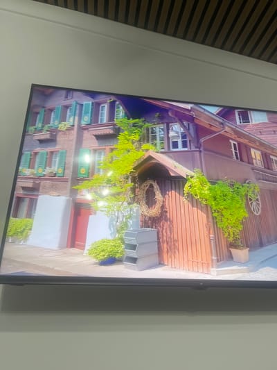 This image depicts a television screen displaying a scenic view of traditional European architecture, possibly in a village setting. The building in the image features dark wood siding, green shutters on windows, and a rustic charm with decorative elements like a wreath and a wooden wheel on the exterior. Lush green foliage cascades down the side of the building, suggesting a well-maintained outdoor space. The scene is illuminated by natural light, indicating it is daytime. There is no indication in the image itself that the location is in Rosenovo, Bulgaria.