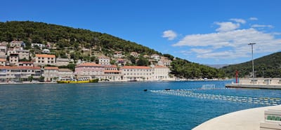 This is a wide-angle, eye-level photograph of a harbor in Općina Pučišća, Croatia. The image is dominated by the vibrant blue water of the sea in the foreground, which occupies the lower half of the frame. A concrete promenade curves into the lower right corner of the image, with white bench seating visible. In the mid-ground, a yellow ferry is docked, its deck lined with passengers. Behind the ferry, a row of multi-story buildings with terracotta roofs line the waterfront. The buildings are a mix of architectural styles, with some featuring stone facades and others painted in pale colors. Behind the buildings, a densely forested hillside rises steeply, covered in dark green pine trees. The sky is a clear, bright blue with a few wispy white clouds scattered across the upper portion of the frame. A tall lamppost stands on the right side of the harbor, near a red marker buoy and a floating water polo goal. The overall impression is one of a sunny, idyllic coastal town on a clear summer day.