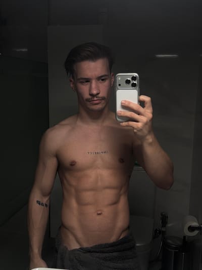 A shirtless man takes a selfie in a bathroom mirror. He has dark hair styled back, a mustache, and several tattoos. One tattoo is vertically written on his chest, spelling out "IMPERFECT". He holds a phone in his right hand. A dark towel is wrapped around his waist. A toilet and a roll of toilet paper are visible in the background. The bathroom has dark lighting.