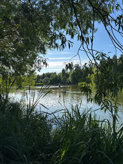 The image depicts a tranquil lake scene in Sloboda Staraya, Russia, framed by lush green foliage. In the foreground, tall grass and reeds line the water's edge. The middle ground reveals a calm lake with gentle ripples, reflecting the blue sky dotted with wispy white clouds. Two individuals are visible on the water: one person is standing on a paddleboard, holding a paddle, while another person is in the water, appearing to be on a similar board, with only their head visible above the surface. In the background, a dense forest of trees lines the far bank of the lake, creating a natural, serene backdrop. The overall impression is one of a peaceful, sunny day spent enjoying outdoor recreation on the water.