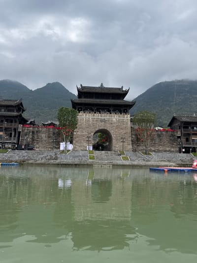 The image depicts a traditional Chinese architectural scene located in Zhuoshui, China. A stone archway, possibly a city gate, dominates the center of the frame. The archway is flanked by traditional wooden buildings with ornate roofs. A mountain range rises in the background, shrouded in mist. A body of water, likely a river or lake, is in the foreground, reflecting the buildings and archway. Small trees are planted near the archway, adorned with red decorations. The sky is overcast, contributing to a subdued and atmospheric feel.