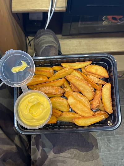 A close-up overhead shot shows a black plastic container filled with golden-brown potato wedges, some with a light dusting of seasoning. To the left, a clear plastic dipping container is open, with a small dollop of yellow sauce on the lid and a generous amount of sauce within the container. The food is placed on a surface with camouflage-patterned fabric underneath. In the background, out of focus, a wooden surface and electronic equipment are visible. The context of Shiryaevo, Russia, is not evident in the image.