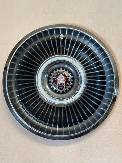 A vintage Mercury Cougar hubcap is mounted on a light beige, textured wall. The hubcap is circular, with a silver outer rim and numerous dark, ribbed spokes radiating towards the center. In the center of the hubcap is a smaller, circular emblem featuring a red background with a silver and red crest depicting a leaping cougar, a crown, and a sword. The words "MERCURY COUGAR" are visible in a circular text around the emblem. The hubcap appears to be made of metal and shows some signs of wear. The lighting suggests an indoor environment. The image does not provide any information about its location in Graham, United States of America.