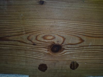 The image is a close-up, overhead shot of a wooden surface. The wood grain is prominent, with horizontal lines running across the frame, interspersed with darker, irregular patterns indicative of knots. There are at least three circular indentations visible, two towards the bottom left and one towards the bottom right, with one on the left appearing darker and more pronounced. The surface appears weathered and aged, with a slightly rough texture. There is no indication of any specific location in Merenii Noi, Moldova, nor are there any people, actions, or interactions depicted.