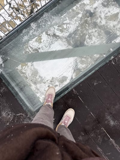 The image displays a top-down perspective of a person standing on a glass-bottomed walkway or bridge in Мастоўскі сельскі Савет, Belarus. The individual's lower legs are visible, wearing grey trousers and light-colored sneakers with pink laces. A dark brown garment is visible in the foreground. The clear glass panel directly beneath the feet is partially obscured by patches of snow, ice, and water droplets. Below the glass, a turbulent, muddy-brown body of water with white foam and ice is discernible. To the left of the glass section, a metal grate structure is visible over the water. To the right, the adjacent walkway consists of dark wooden planks, also bearing scattered snow and ice.