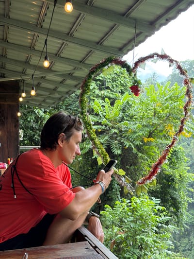 A man in a coral-colored shirt stands with his head tilted upward, holding a black smartphone to take a picture of a heart-shaped decoration made of greenery and red tinsel. The decoration hangs against a backdrop of lush green foliage. A string of warm-toned lights runs along a corrugated fence on the left. The man has short brown hair, is wearing bracelets, and is partially obscured by a wooden structure.