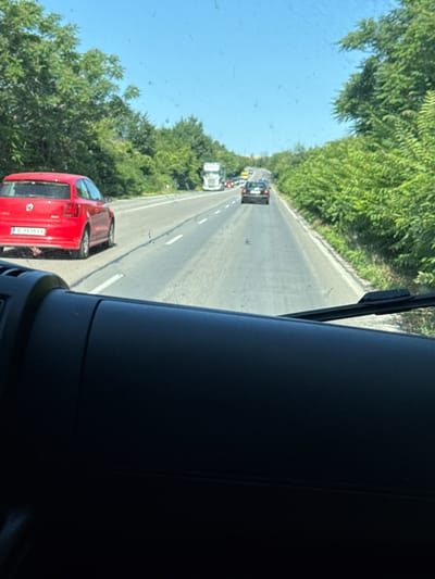 A red Volkswagen Polo is driving on a highway with dense green trees lining the sides. The car is in the left lane, and there are other vehicles on the road ahead, including a white truck and a dark-colored car. The sky is clear and blue. The image is taken from inside another vehicle, likely a car, as indicated by the dashboard visible in the foreground. The license plate of the red car appears to be from Bulgaria. The location is identified as the city of Osenets, Bulgaria.