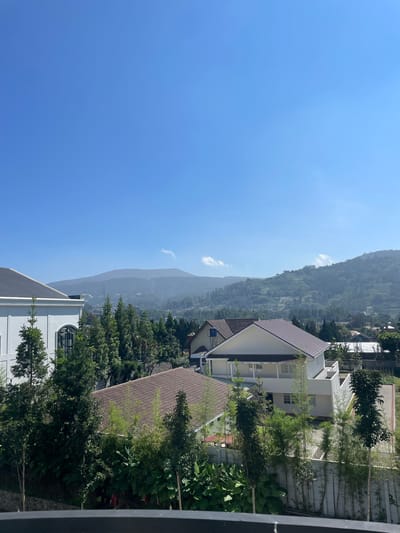 This image depicts a daytime panoramic view over the city of Kayuambon, Indonesia, showcasing a residential area set against a mountainous backdrop under a clear blue sky. The sky occupies approximately the upper third of the frame, exhibiting a vibrant blue hue with a few small, wispy white clouds visible, primarily towards the center-right horizon.

In the mid-ground, a range of verdant mountains dominates the horizon, appearing hazy due to atmospheric conditions. The mountain slopes are densely covered with foliage, and some areas show distinct terraced patterns, indicative of cultivation. Distant structures, possibly communication towers, are faintly discernible on the highest peaks.

The foreground and mid-ground are populated by several residential buildings interspersed with lush greenery. On the far left, a large, modern white building with a dark grey tiled roof is partially visible. Its design features large windows and arched details. To its right, a dense cluster of trees, including tall conifers and various leafy species, obscures parts of the landscape.

Towards the right side of the frame, another prominent house is visible, characterized by white walls and a warm brown tiled roof. This house appears to have multiple levels and balconies. Further right, a concrete or stone retaining wall with vertical bamboo-like plants is present, suggesting landscaping or boundary demarcation. Various other houses and structures with different roof colors (brown, white) are scattered throughout the mid-ground, nestled among abundant trees and foliage. A water tank is visible on the roof of a house towards the left-center, above the main cluster of trees. No people are visible in the scene. The lighting suggests a bright, sunny day, with clear shadows.