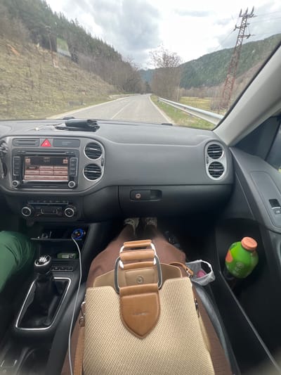 The image presents a first-person view from the front passenger seat of a vehicle, identified as a Volkswagen Tiguan based on interior design, navigating a paved road in the mountainous region of Cherna Mesta, Bulgaria. The time displayed on the car's infotainment system is 12:51.
The exterior environment depicts an overcast sky above a two-lane asphalt road that curves gently to the right in the distance. On the left, a steep, forested hillside rises, covered with mostly bare deciduous trees and some scattered evergreens. On the right, a white metal guardrail parallels the road, beyond which lies a grassy verge and another forested incline, featuring a prominent red metal power line tower with multiple insulators and wires.
Inside the vehicle, the dashboard is grey-black. The center console includes an infotainment system with "ENERGY" displayed and the time "12:51," along with climate controls below it. A red hazard warning light button is illuminated. On the driver's side, a person's leg, clad in green trousers, is partially visible next to a 6-speed manual gear shifter. A USB charger with a blue indicator light is plugged into a compartment, with a white cable extending from it. On the passenger's side, the legs and feet of a person wearing brown trousers and light-colored sneakers are visible. A tan and brown Burberry handbag with a prominent silver-colored buckle rests on the passenger's lap. In the passenger door's storage pocket, a green plastic bottle with an orange cap and a small white plastic bag containing a pink object are present. No direct actions or interactions between individuals are depicted, beyond their presence in the vehicle.