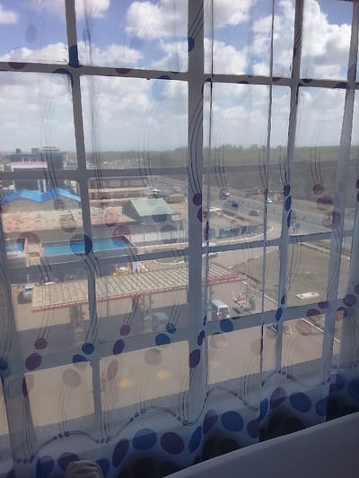 The image displays an elevated view of an urban landscape in Gatongora ward, Kenya, observed through a multi-paned window. A translucent white curtain, decorated with blue, purple, and red circular patterns and wavy lines, partially obscures the scene.
Through the window, a Shell Energies gas station occupies the mid-ground, characterized by a red and white canopy structure, multiple visible fuel pumps, and several vehicles, including a light-colored van, positioned at the pumps. Adjacent to and beyond the gas station, various low-rise commercial or industrial buildings with distinct roof colors (blue, green, and brown) are visible. A curved multi-lane road or highway traverses the scene, with several vehicles in transit. The background consists of a distant tree line or dense vegetation under a partly cloudy blue sky with scattered white clouds. No specific individuals are clearly identifiable.