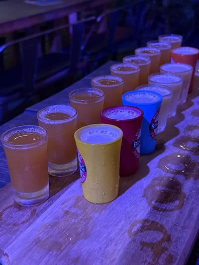 An array of at least fifteen small beverage vessels is positioned in a linear formation on a light-colored wooden surface within a dimly lit indoor environment, likely a bar or brewery in Noumea, France. The scene is illuminated by a strong purple-blue ambient light. The foreground displays a sequence of vessels: clear glass shot glasses containing an amber liquid with foam, interspersed with three opaque plastic cups. From left to right, these plastic cups are yellow, red, and blue, each featuring partial branding that includes the text 'BING PILSENER BEER'. The yellow cup exhibits external condensation droplets. The wooden surface, resembling a serving paddle, is marked with numerous distinct circular wet rings and larger damp patches. The background is out of focus, showing indistinct forms of furniture under the same low, purple-blue illumination. No individuals are present.