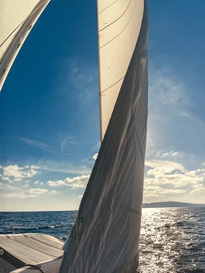 The image is a vertical, low-angle shot from the perspective of someone on a sailboat. Two white sails are prominently featured, one on the left and another larger one dominating the right side of the frame. The sail on the right is catching the sunlight and appears to be billowing. The sky is a vibrant blue with scattered white clouds. The sea is choppy with sunlight glinting off the waves. In the distance, a hazy landmass, possibly an island or coastline, is visible on the horizon. The foreground shows part of the sailboat's deck, including some padded seating. There are no people visible in the image. The scene depicts a bright, sunny day at sea. This image is not of Ossenzijl, Netherlands.