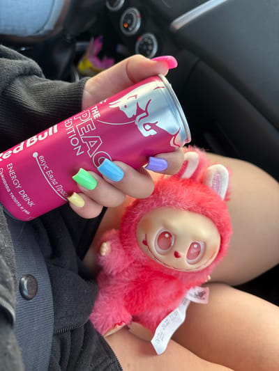 A person is holding a pink can of Red Bull and a fluffy pink toy creature inside a car. The person's nails are painted in pastel colors: neon pink, light blue, lime green, yellow, and lavender. The Red Bull can is labeled "The Peach Edition" and has text in Bulgarian. The toy creature is pink and furry with large eyes and rabbit-like ears. The background shows parts of the car's interior, including the dashboard and seats. The image is a close-up shot, with the focus on the Red Bull can, the toy, and the person's hand. The location in the city of Δρυόφυτο, Greece, is not discernible from the image.