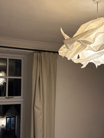 The image presents an interior view under low lighting conditions. Dominating the upper left is an illuminated, stylized paper lantern hanging from the wall. The walls appear to be off-white or light beige. Below the lantern, a dark metal rod extends downwards. To the right, a beige roman blind is positioned above a white-framed window. The window has a multi-pane design, and through it, a dark, indistinct background is visible, possibly an outdoor scene at night. No people are visible in the shot. The location is impossible to determine based on the image.