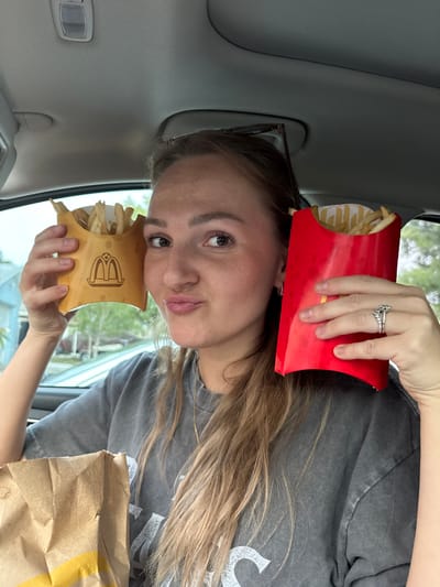 In a vehicle, a young Caucasian woman with blonde hair, wearing a gray t-shirt, poses for a selfie. She is holding two containers of french fries, one in a red container and the other in a gold-colored container with a McDonald's logo. The woman is in the driver's seat, and the background shows a residential area with houses and trees. The image appears to have been taken during the daytime.