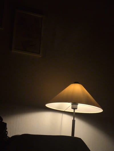 This media file captures a dimly lit interior scene. The primary subject is a cone-shaped lampshade, illuminated from within, casting a warm yellow light onto a textured wall. The lamp is mounted on a metal arm. To the left of the lamp, a framed picture hangs on the wall, its contents obscured by shadow. The overall atmosphere is subdued and quiet, suggesting a nighttime setting. The scene is dark, with the lamp being the dominant light source, creating strong contrasts between illuminated and shadowed areas. There is no visible text. The location context provided is Słupsk, Poland.