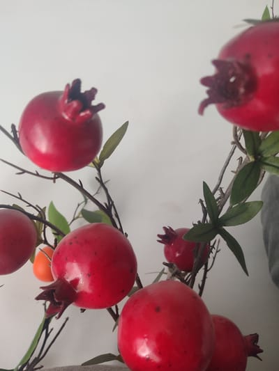 The image shows a close-up of a decorative artificial pomegranate branch. Several bright red pomegranates are visible, attached to thin brown stems with small green leaves. Some of the pomegranates have a darker red, crown-like top. In the background, the wall is a plain, light color, possibly white or off-white, and a grey textured surface is visible on the right side. There are no people or animals in the image. The lighting is soft, suggesting an indoor setting with diffused light. The focus is on the pomegranates, with some of the background slightly blurred. The scene does not contain any specific indicators of its location in Batken City, Kyrgyzstan.
